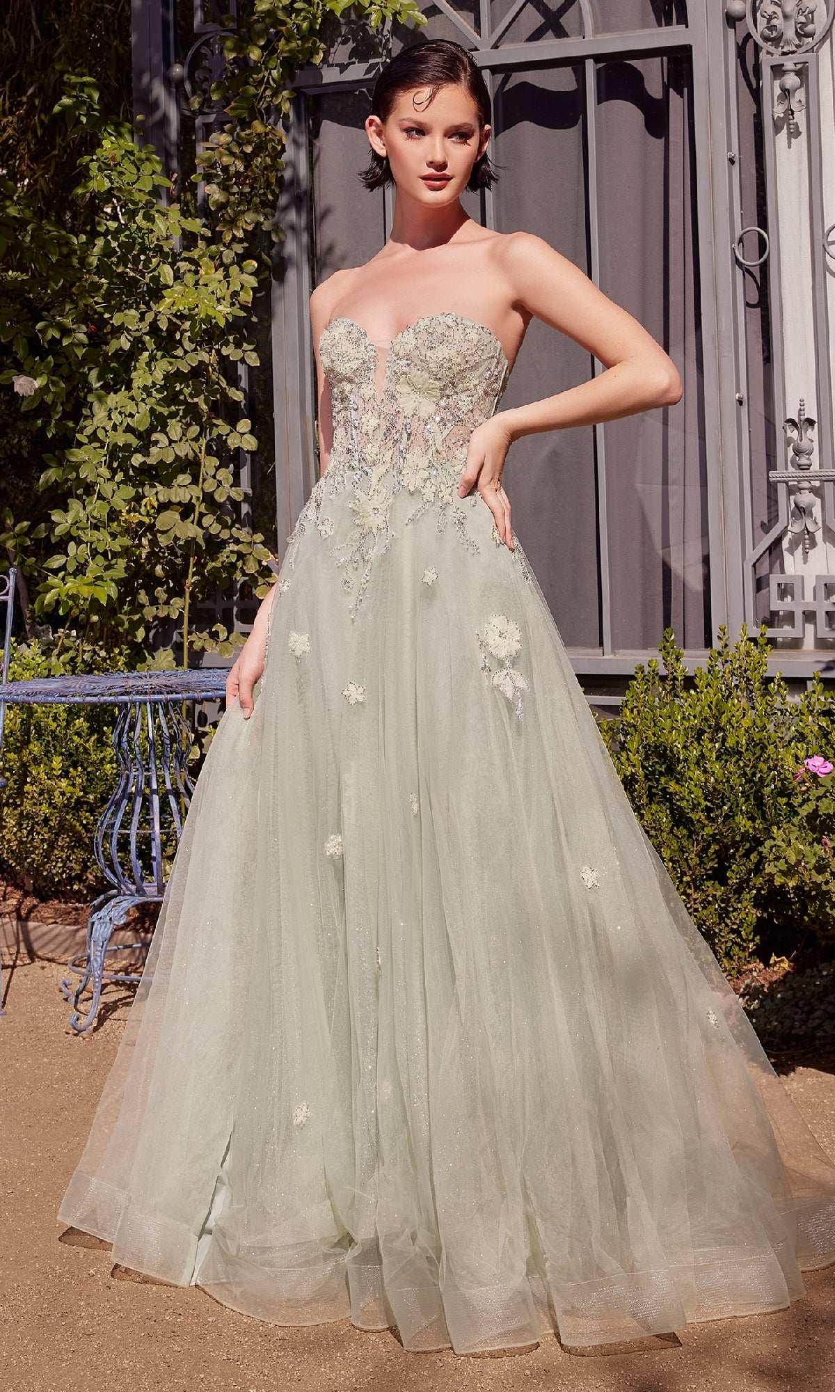 A woman stands outdoors on a gravel path near lush greenery and an ornate wrought iron window, wearing the Sage Green Prom Ball Gown: Andrea & Leo A1418, a strapless corset gown with floral embroidery.